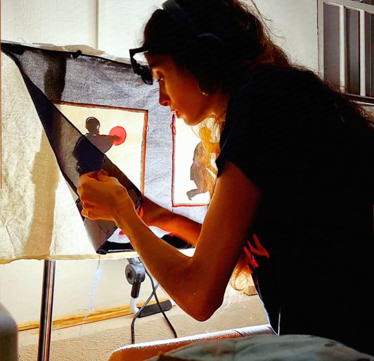 Inès Ré à l'atelier, penchée sur une découpe textile devant une fenêtre à contre-jour.