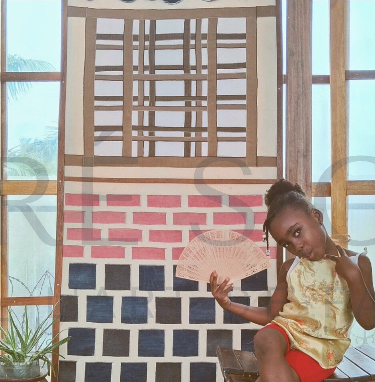 Une enfant tient un éventail devant HANOKA, panneau textile mural aux briques roses et bleues.