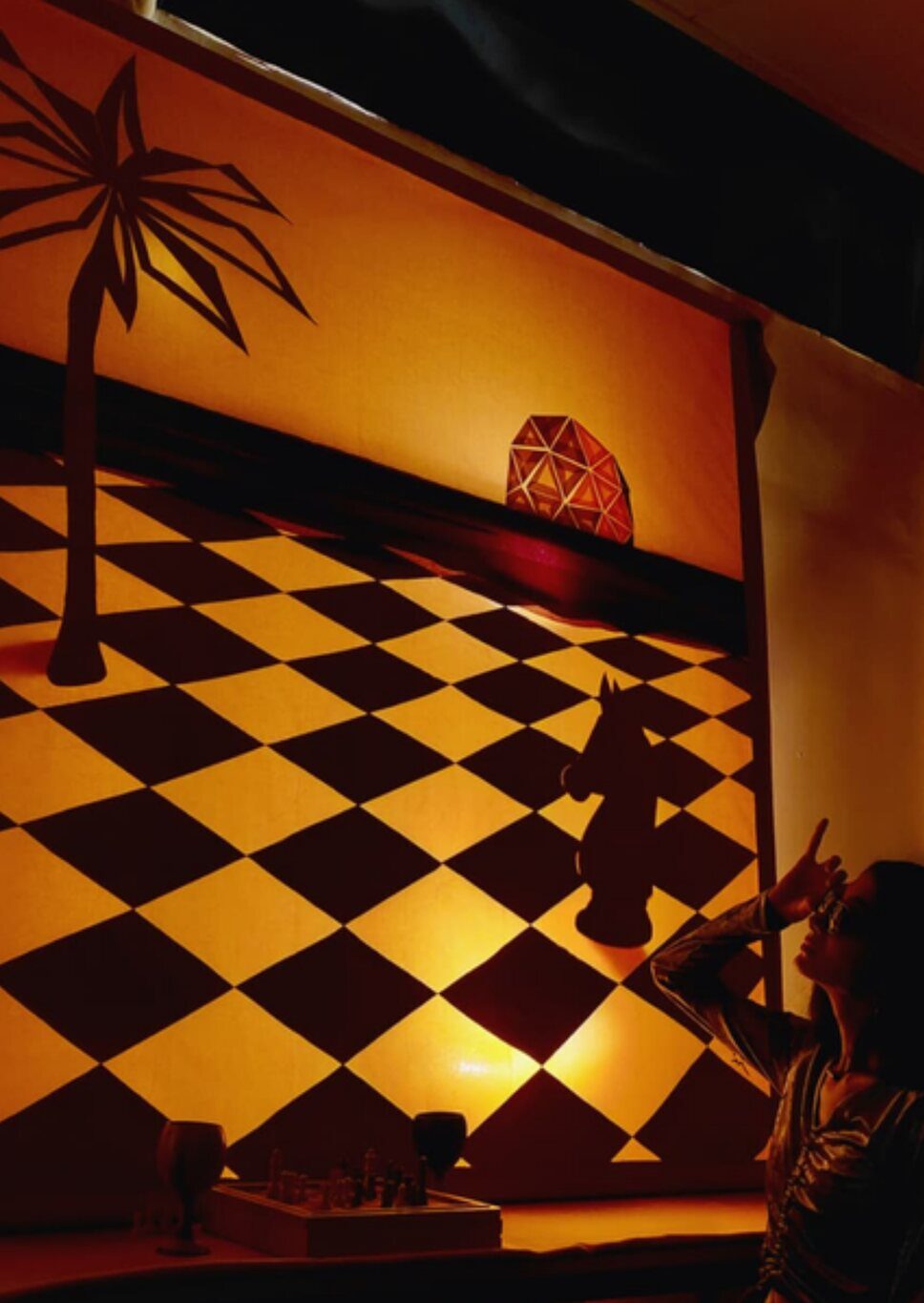 Intérieur tamisé baigné d'une lumière orange, sol en damier noir et or et palmier en ombre chinoise.